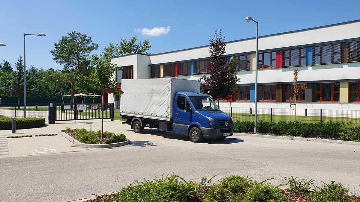 Kék ponyvás kisteherautó érkezik egy modern épülethez kültéri kiszállításhoz